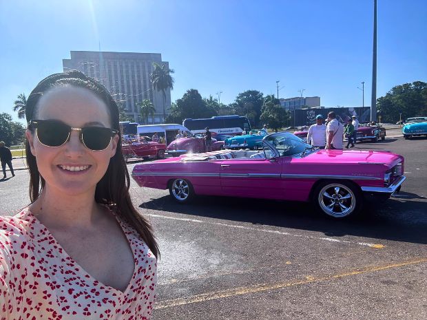 A woman in sunglasses smiles in the foreground while a pink convertible is parked in a sunny outdoor lot. Several people and colorful cars are visible in the background.