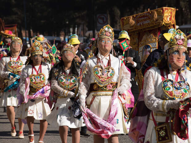 People dressed in ornate, colorful costumes and masks, walking in a festive procession. They wear intricate headpieces and hold decorative items. The background shows other participants and spectators in an outdoor setting.