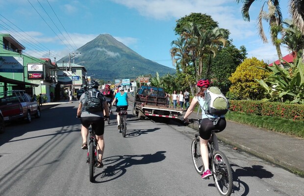 Cyclists ride down a street, flanked by colorful buildings and lush greenery. In the distance, Arenal Volcano towers under a clear blue sky, creating a scenic tropical backdrop.