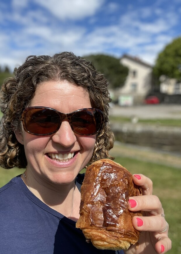 A person wearing sunglasses holds a chocolate croissant, smiling outdoors. A grassy area and distant building under a partly cloudy sky are visible in the background.