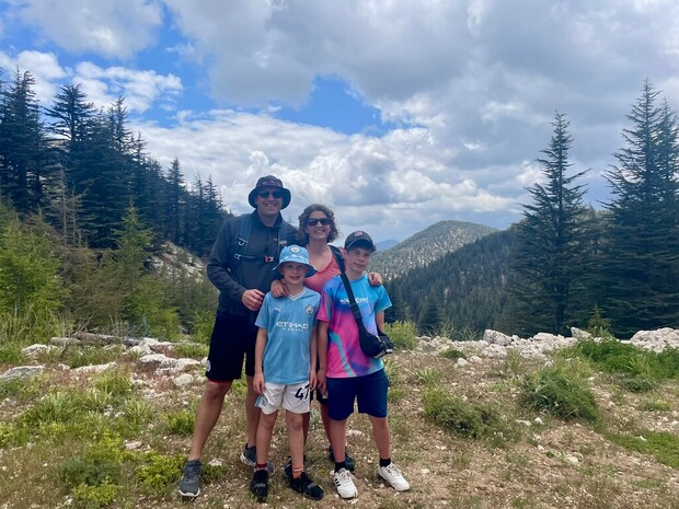A family of four stands smiling on a grassy, rocky hilltop surrounded by tall pine trees and distant, cloudy mountains under a partly cloudy sky.