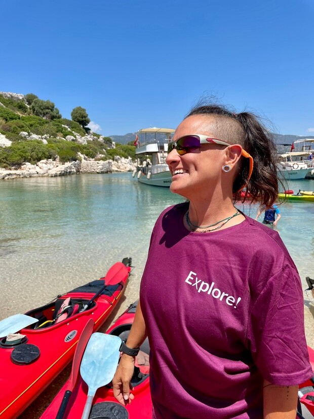 A person wearing sunglasses and a purple "Explore!" shirt stands smiling beside red kayaks near a calm, clear body of water, with boats and rocky foliage in the background.