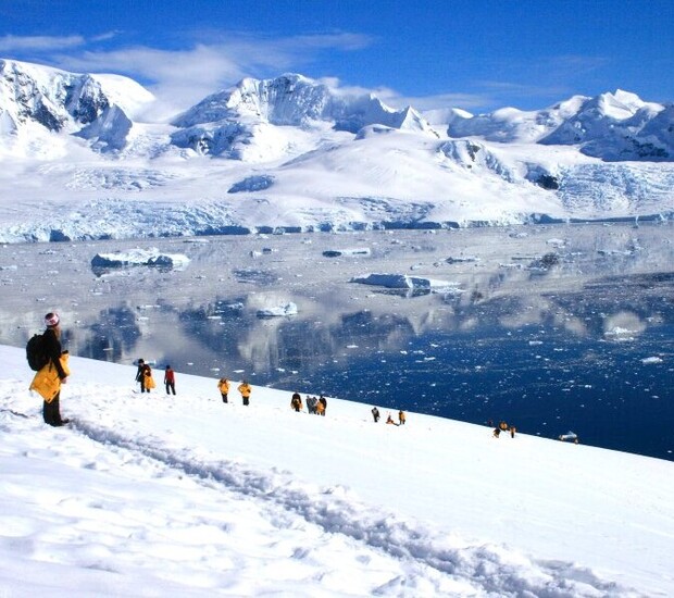 People trek up a snowy slope wearing yellow and black jackets. Icebergs and a serene blue waterbody appear in the background, framed by majestic snow-covered mountains under a clear sky.