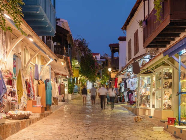 Evening market scene with clothing and souvenir shops on both sides of a cobbled street. People stroll under warm lights with displays of colorful garments and decor items, surrounded by traditional architecture.