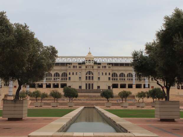 A large, historic stadium stands silently in focus, flanked by trees. A rectangular reflecting pool stretches in the foreground, enhancing the symmetry of the tranquil, overcast setting.