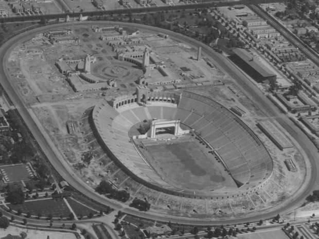 Stadium with a large oval-shaped seating area surrounds a grass field, viewed from above. Construction work is visible around, with urban buildings in the background, indicating a developing city.