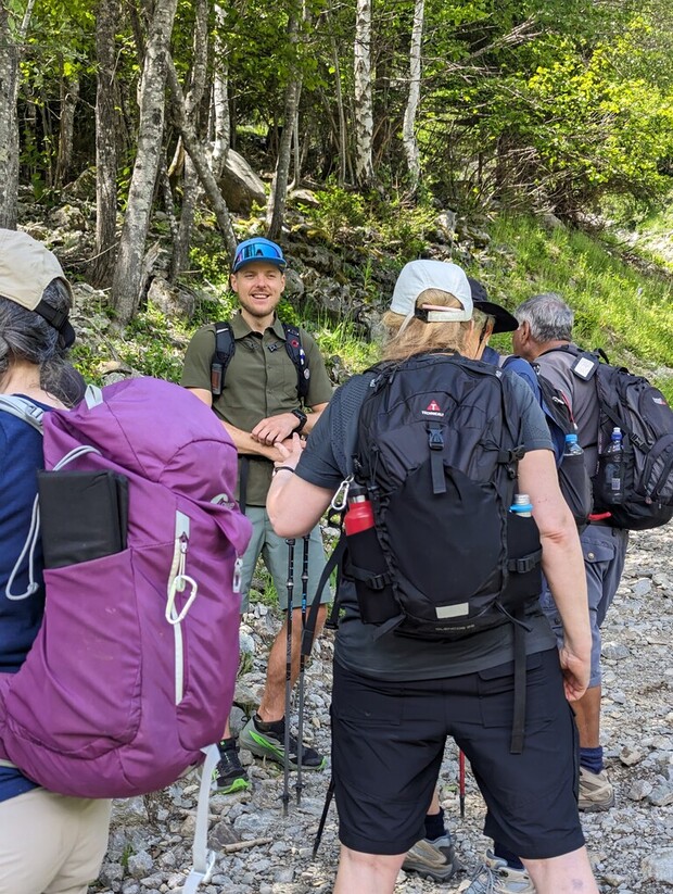 Hikers with backpacks converse on a rocky trail in a forested area, surrounded by green foliage and trees. One person faces the group, appearing to address them.