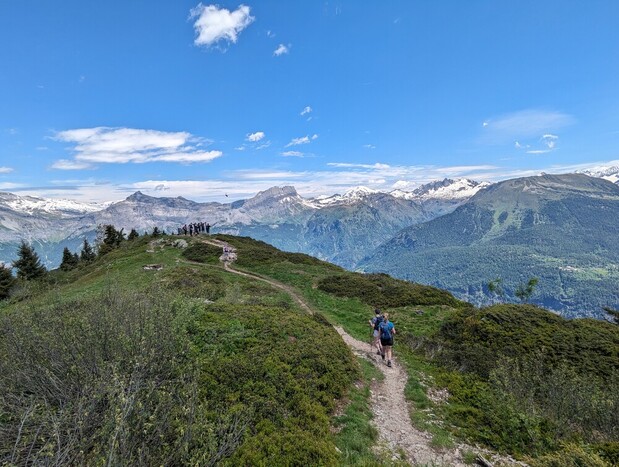A hiker walks along a narrow, winding trail on a grassy ridge, surrounded by snow-capped mountains and a clear blue sky. Others gather in the distance.