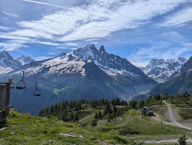 Snow-capped mountain peaks tower under a blue sky with wispy clouds, overlooking a verdant valley. A ski lift with empty chairs stretches across the scene, enhancing the tranquil alpine setting.