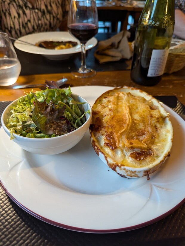 A baked dish and salad sit on a white plate on a restaurant table. Nearby, there are glasses of red wine and water, with another dish visible in the background.