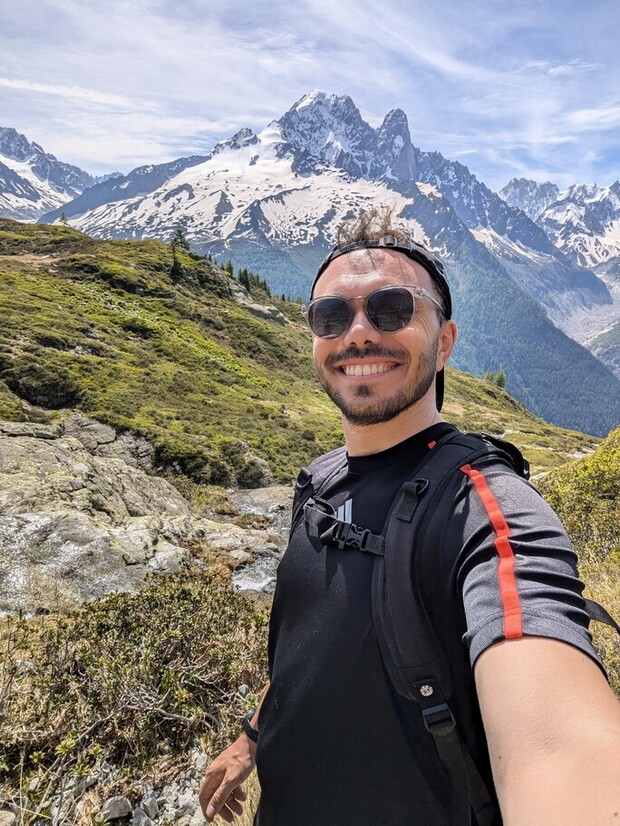 A person wearing sunglasses and a backpack smiles while taking a selfie. They stand in a mountainous area, surrounded by green hills and snow-capped peaks under a clear sky.