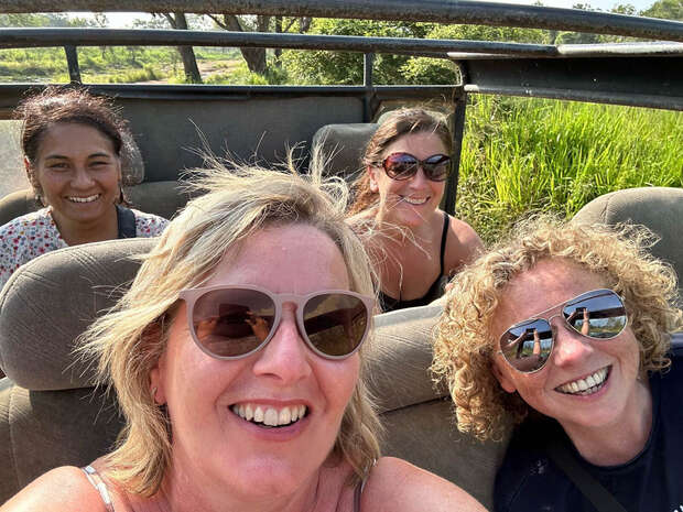 Four people smile while sitting in an open-top vehicle, surrounded by lush greenery under a clear sky, suggesting a safari or nature excursion in a sunny, outdoor environment.