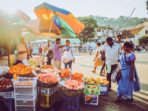 Fruit vendor stands under a colorful umbrella, selling various fruits displayed in baskets and crates on a busy street. People are around, some walking or stopping to buy.