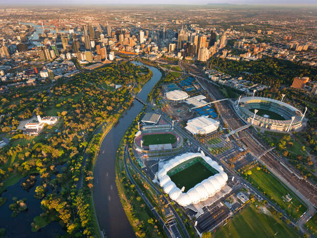 Aerial view of a cityscape featuring a winding river surrounded by parks and sports stadiums, with a densely built urban skyline in the background, under clear skies.