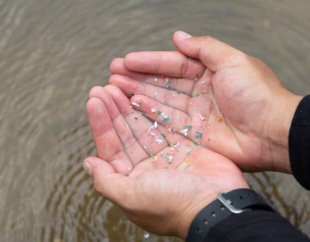 Hands holding colorful microplastic particles above a water surface, creating ripples. A watch is visible on one wrist, indicating casual attire and focusing on environmental issues.