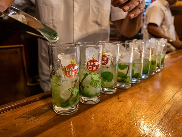 A person pours liquid into glasses labeled "Havana Club," each filled with ice and mint, on a wooden bar counter in a lively setting with another person in the background.