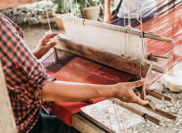Hands operate a traditional wooden loom, weaving red fabric with white threads. The person wears a checkered shirt. Pot plants and another person are visible in the background.