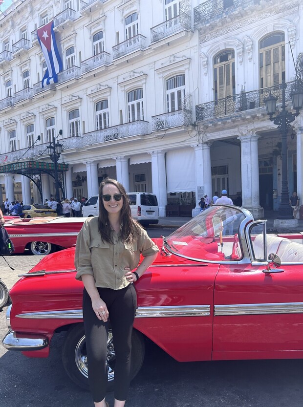A person stands by a red vintage convertible car, smiling, with a white colonial-style building and a Cuban flag in the background, surrounded by people and other classic cars.