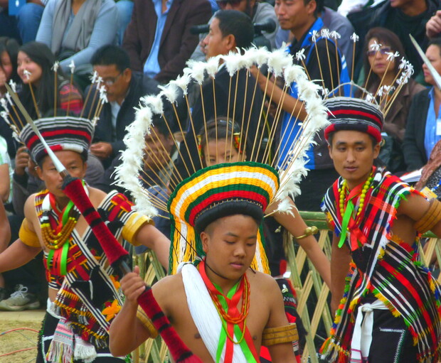 People wearing traditional, colorful attire with feathered headdresses perform a dance. They're in an outdoor setting, surrounded by onlookers. The detailed costumes feature vibrant patterns and accessories.