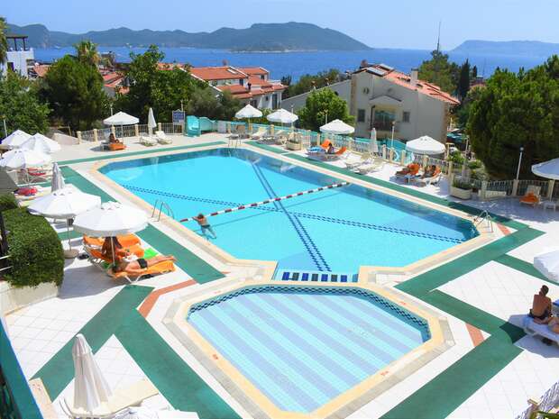 A large outdoor swimming pool with lanes, surrounded by lounge chairs and umbrellas, has a person swimming. It's set in a sunny resort with trees and a view of distant mountains and the sea.