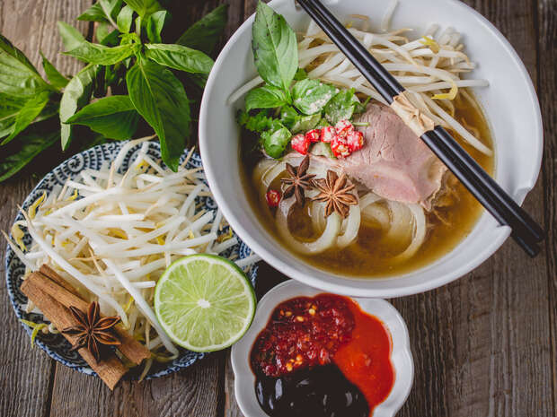 A bowl of noodle soup with sliced meat, herbs, bean sprouts, and star anise, garnished with lime. Accompanied by a dish of red sauces and a blue bowl of extra sprouts, set on a wooden table.