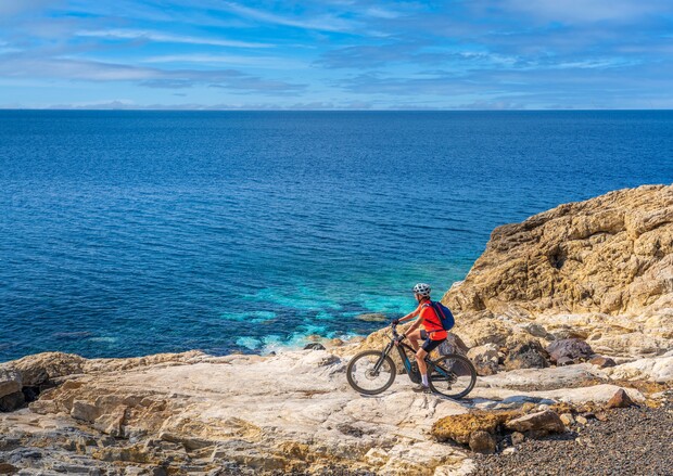 A cyclist wearing a helmet and backpack rides a bike on rocky terrain overlooking a vast blue ocean under a clear, partly cloudy sky.