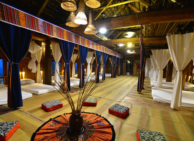 A traditional room with futon-style beds aligned under colorful fabric canopies, wooden beams overhead, woven mats, and decorative pillows, creating a serene and cultural atmosphere.