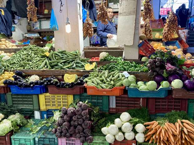 Vegetables are stacked in colorful plastic crates at a bustling market. A vendor stands among hanging produce, surrounded by cucumbers, eggplants, carrots, and cabbage, under bright lighting.