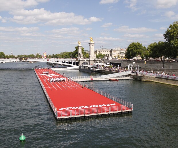 A floating red track with "#PARIS2024" hosts people gathering, positioned on a river near a bridge with golden statues, surrounded by crowds and trees under a partly cloudy sky.