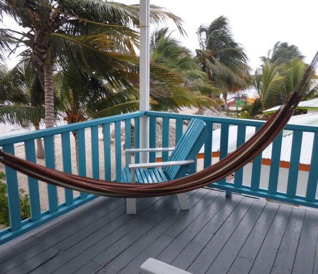 A hammock hangs motionlessly on a wooden balcony, accompanied by a blue Adirondack chair. Palm trees sway in the background, indicating a tropical, windy beachfront setting.