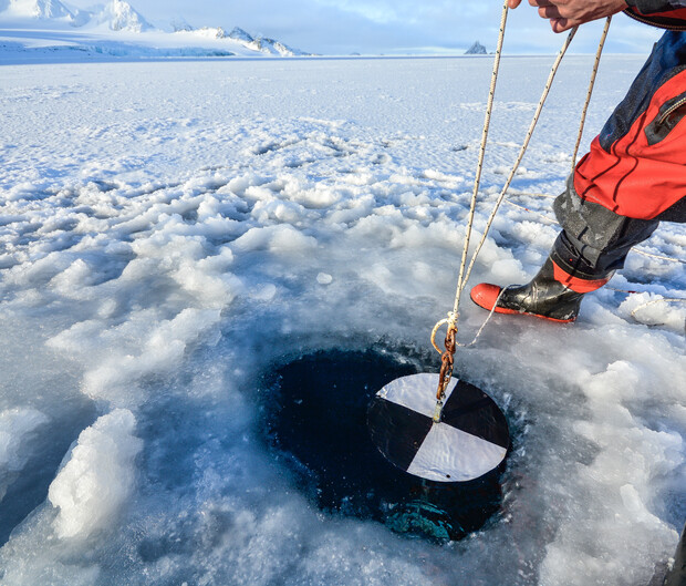 Person lowers a black-and-white disc into an icy hole using a rope. They are standing on a snowy, frozen surface under a clear sky with distant mountains.