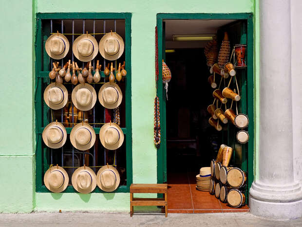 Hats and maracas hang on a green wall, while drums and traditional instruments fill a shop doorway beside them, suggesting a vibrant cultural market setting.