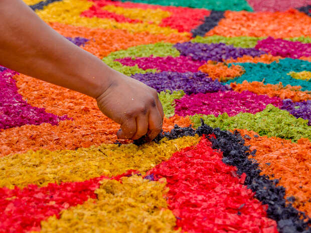 A hand arranges vibrant, multicolored sawdust into intricate patterns on the ground, creating a lively and colorful design as part of an outdoor decorative activity.