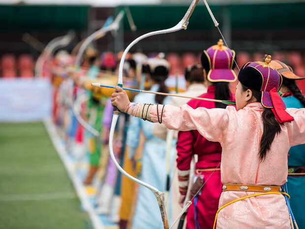 Archers in traditional attire draw bows in unison on a grassy field during a cultural event, with colorful clothing and a focused stance emphasizing precision and coordination.