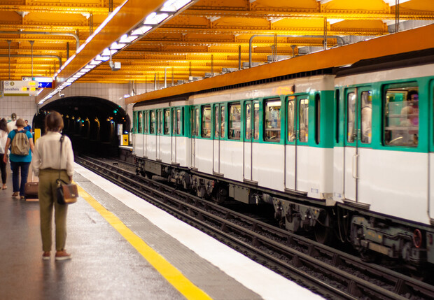 A subway train is arriving at an underground platform, with several passengers waiting. The ceiling is illuminated with yellow lights, and signs indicate directions for "Sortie" and "Correspondances."