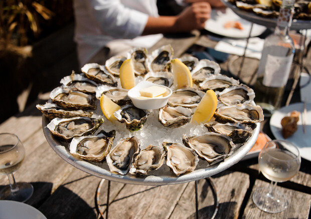 A platter of fresh oysters arranged on ice, garnished with lemon wedges, sits on a rustic wooden table. A person nearby prepares to enjoy a meal, with drinks and plates around.