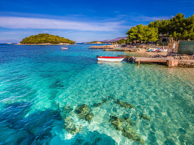 A small boat floats in crystal-clear turquoise water near a stone pier. Lush green trees line the shoreline, with distant islands under a vivid blue sky.