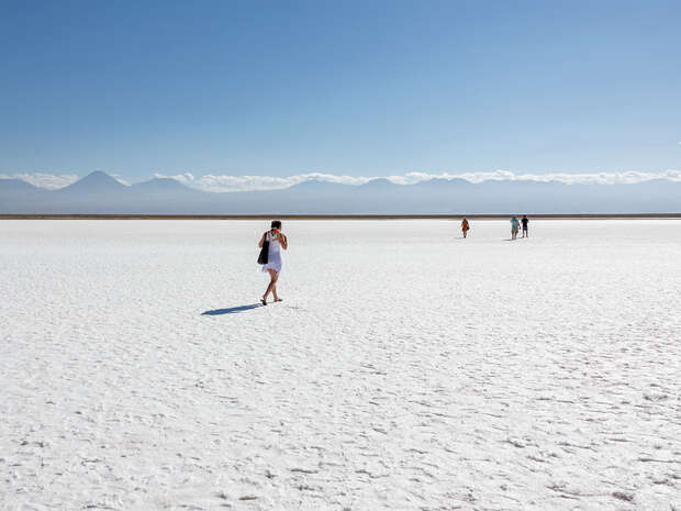 People walk across a vast, white salt flat under a clear blue sky, with distant mountains on the horizon.