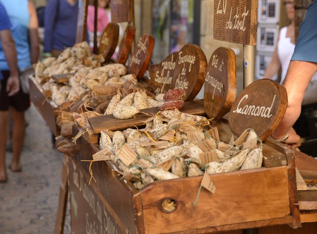 Assorted cured sausages rest in wooden crates, marked with circular signs like "Fromage de Chèvre" and "Canard," at an outdoor market. People walk by on a cobblestone street.