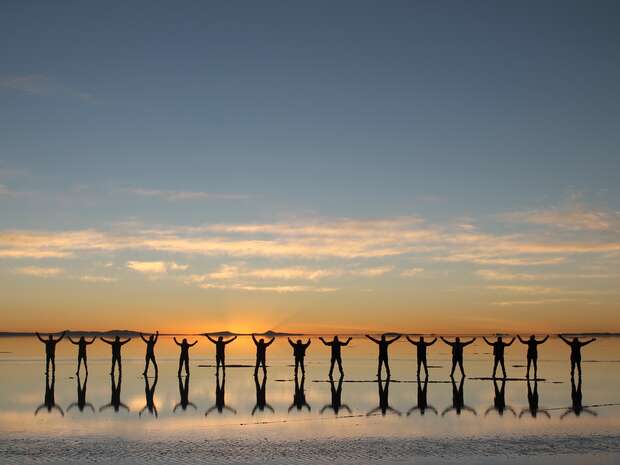 Silhouetted people stand on reflective water, arms raised, against a backdrop of a colorful sunset with scattered clouds in the sky.