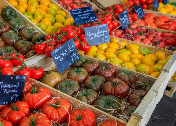 Variety of tomatoes displayed in wooden crates, labeled with chalkboard signs. Text: "FRANCE - Noire de Crimée - €3,99/kg," "Pleine - Tomate grappe Rouge - €1,99/kg," "Perny longue de Picore - €2,99/kg." Background shows similar produce arrangement.