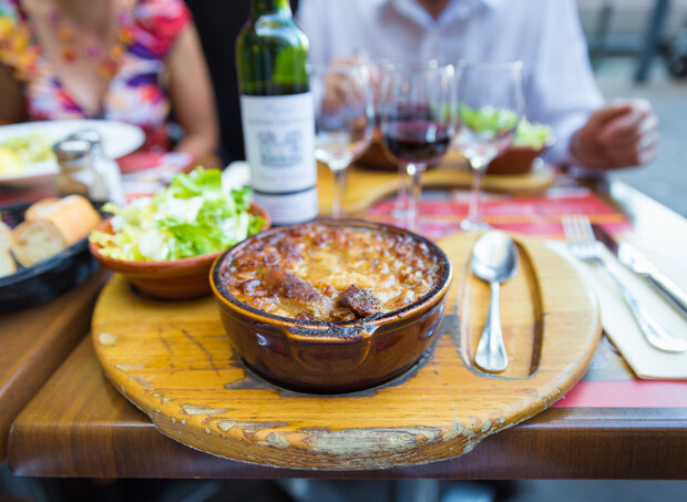 A steaming casserole rests on a wooden board, accompanied by a side salad, wine bottle, and glasses, set on an outdoor restaurant table with diners in the background.
