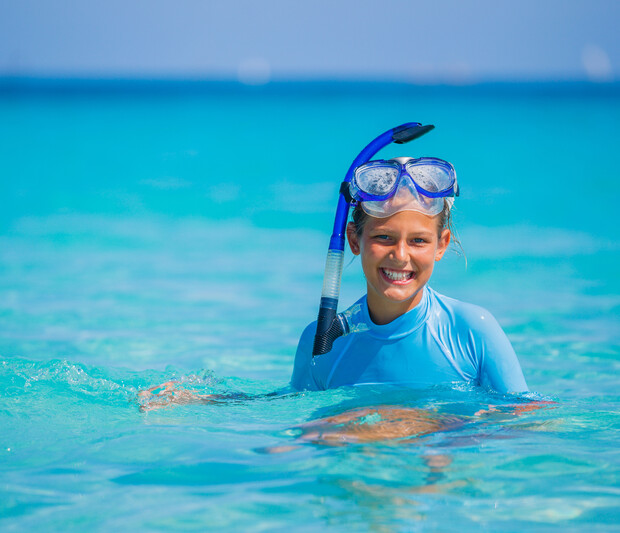 A person wearing a snorkel set smiles while partially submerged in clear turquoise water, with an expansive ocean view under a blue sky in the background.