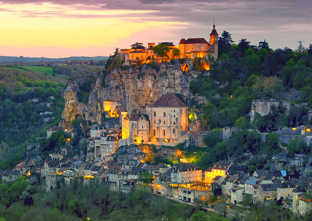 A historic village perches on a rocky cliff at sunset, with illuminated buildings cascading down the hillside, surrounded by lush greenery and a vast landscape stretching into the distance.