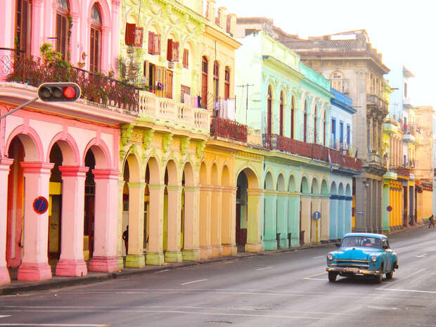 A vintage blue car drives past colorful, arched buildings in various pastel hues, lining a quiet urban street, with a traffic light overhead and clear sky above.