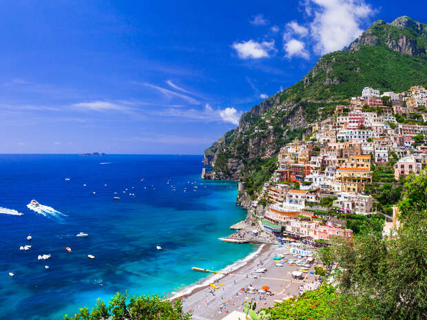 Colorful buildings hug a steep hillside, descending to a busy beach along clear, blue ocean waters dotted with boats under a bright, clear sky.
