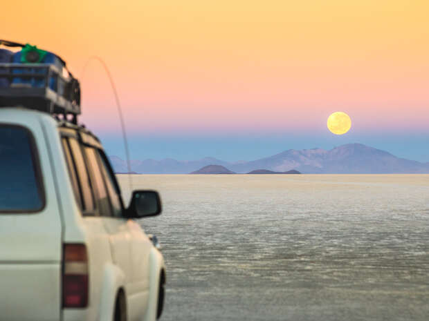A white SUV with roof cargo stands stationary on a vast salt flat at sunset. The full moon rises above distant mountains under a pink and orange sky.