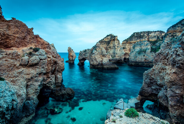 Rock formations arch over turquoise water, surrounded by steep cliffs in a coastal landscape. Steps lead down into the water, and the sky is overcast, creating a serene atmosphere.