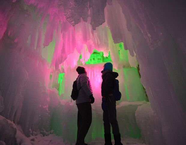 Two people stand inside an ice cave, illuminated by vibrant pink and green lights, surrounded by thick, jagged ice formations. One person wears a blue beanie, while the other has a backpack.