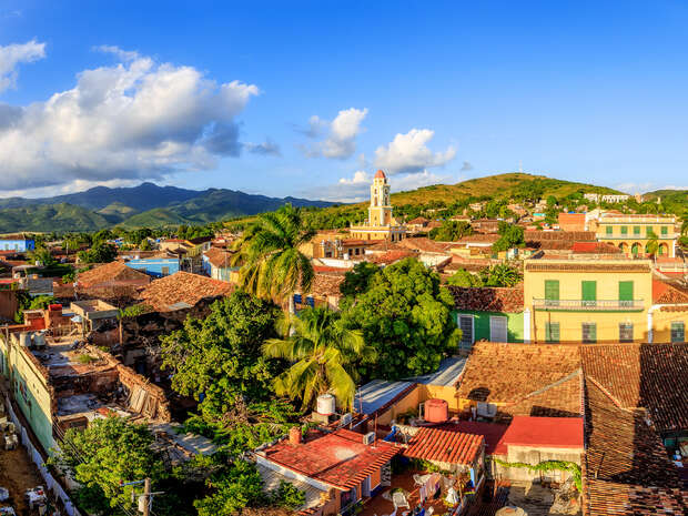 Colorful buildings with red-tiled roofs stand amidst lush greenery. A tall, yellow clock tower rises in the background, under a clear blue sky with scattered clouds and distant mountains.